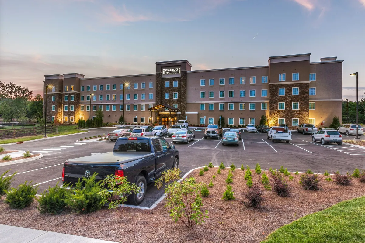 This image showcases the hotel's entrance, with a beautifully landscaped parking lot, stone façade, and large inviting windows. The view captures the welcoming ambiance as guests arrive, set against a soft sunset, suggesting proximity to shopping and dining adventures just outside.