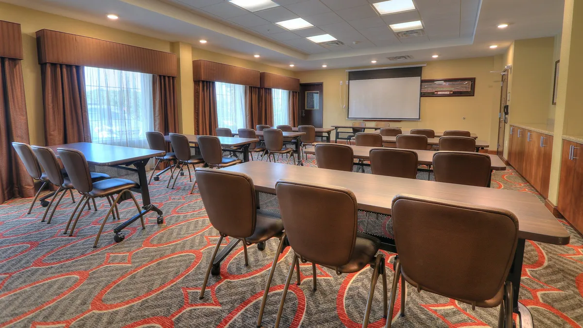 The image depicts an interior meeting space arranged with tables and chairs facing a projector screen. The room is illuminated with natural light filtering through the curtains, creating a professional yet comfortable setting. This environment is designed for business meetings, seminars, or collaborative work sessions, with features that support focused productivity and group interaction.
