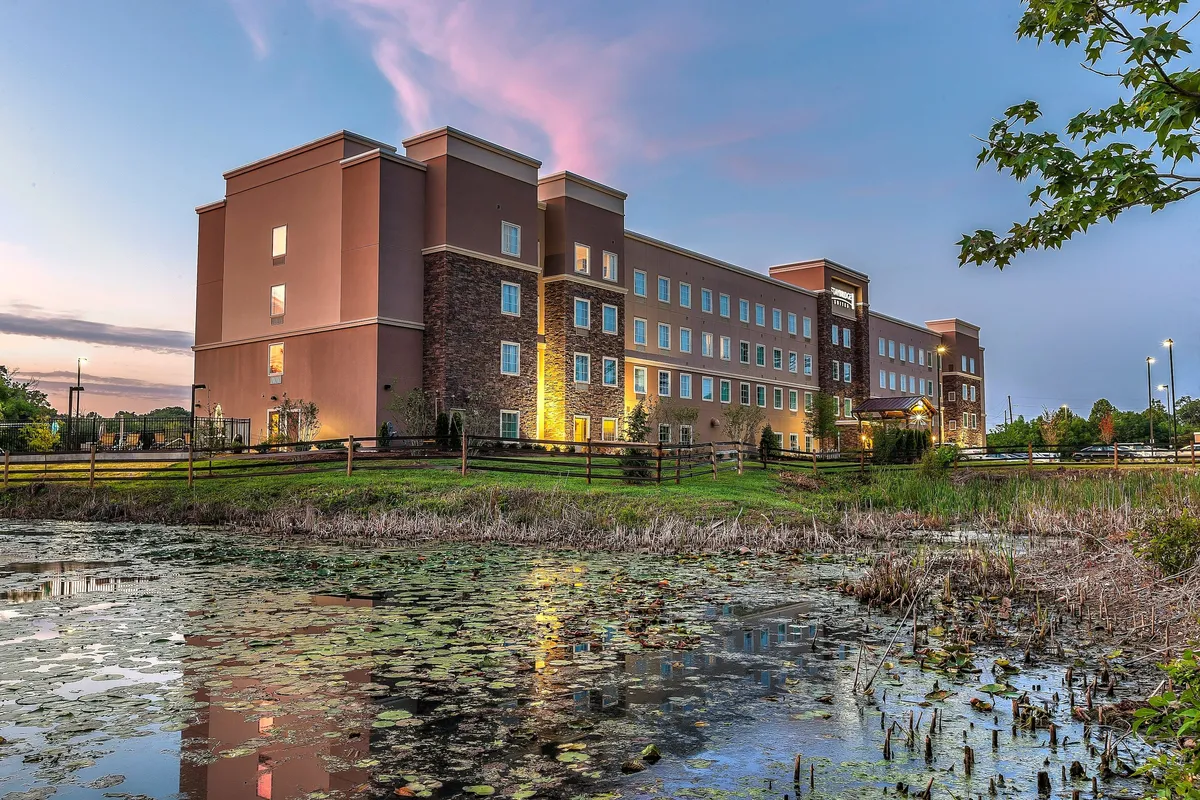 A serene pond bordered by lush greenery, with the hotel in the background. The scene evokes relaxation and outdoor enjoyment, showing guests strolling by the water or pausing for sunset views, hinting at the area's inviting surroundings and walkability.