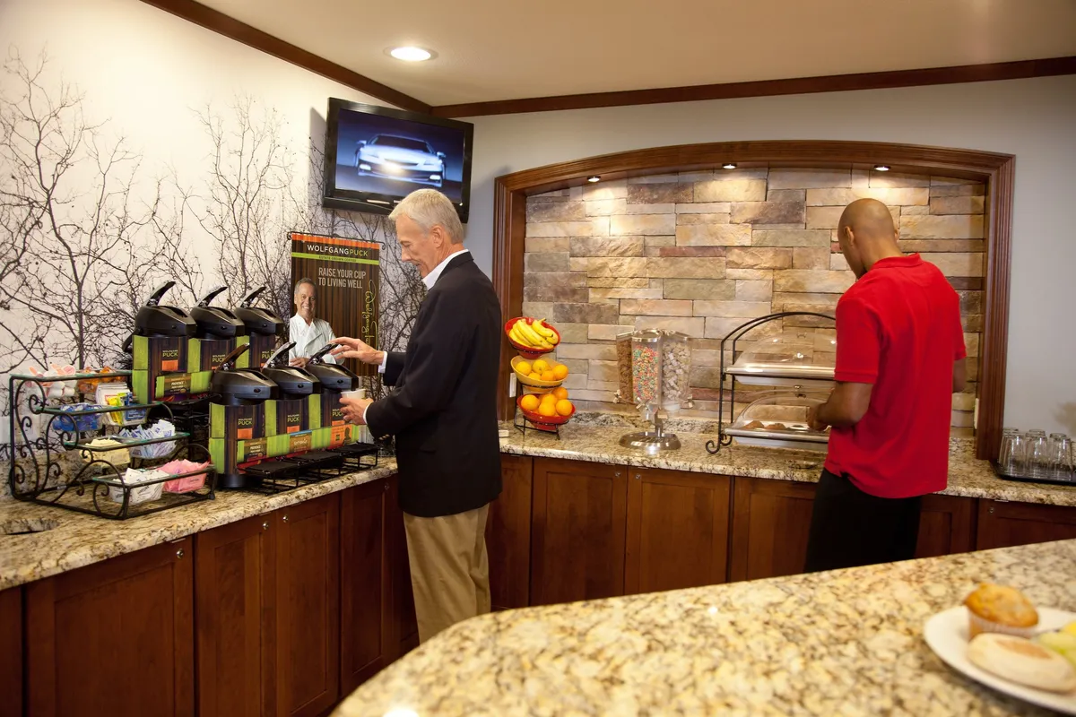 A bright, communal breakfast area featuring coffee dispensers, a fresh fruit display, and a sleek granite countertop. People are gathered, socializing over breakfast and enjoying coffee in a relaxed atmosphere. Artistic wall décor adds a welcoming touch.