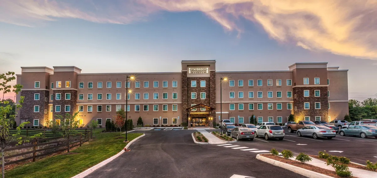 This exterior image showcases the hotel's welcoming facade, framed by lush landscaping and highlighted by an inviting entrance awning. The scene is captured at sunset, adding a tranquil and warm atmosphere. Visible are the parking area and outdoor spaces, giving viewers a sense of arrival and connection to the outdoors.